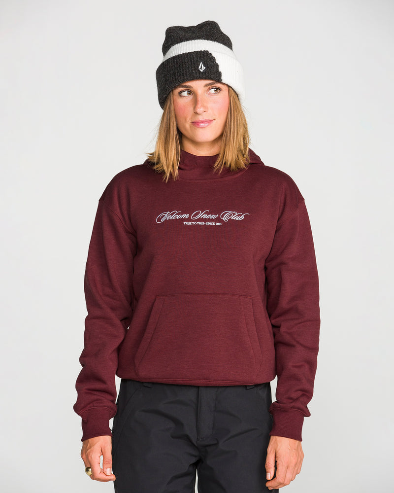 A woman stands against a plain light background, looking slightly to the left, dressed in the Volcom Women’s Alpine All Day Pullover—a maroon hoodie with “Skiers Union Club” text and a cozy handwarmer pocket—paired with black pants and a black and white knit beanie.