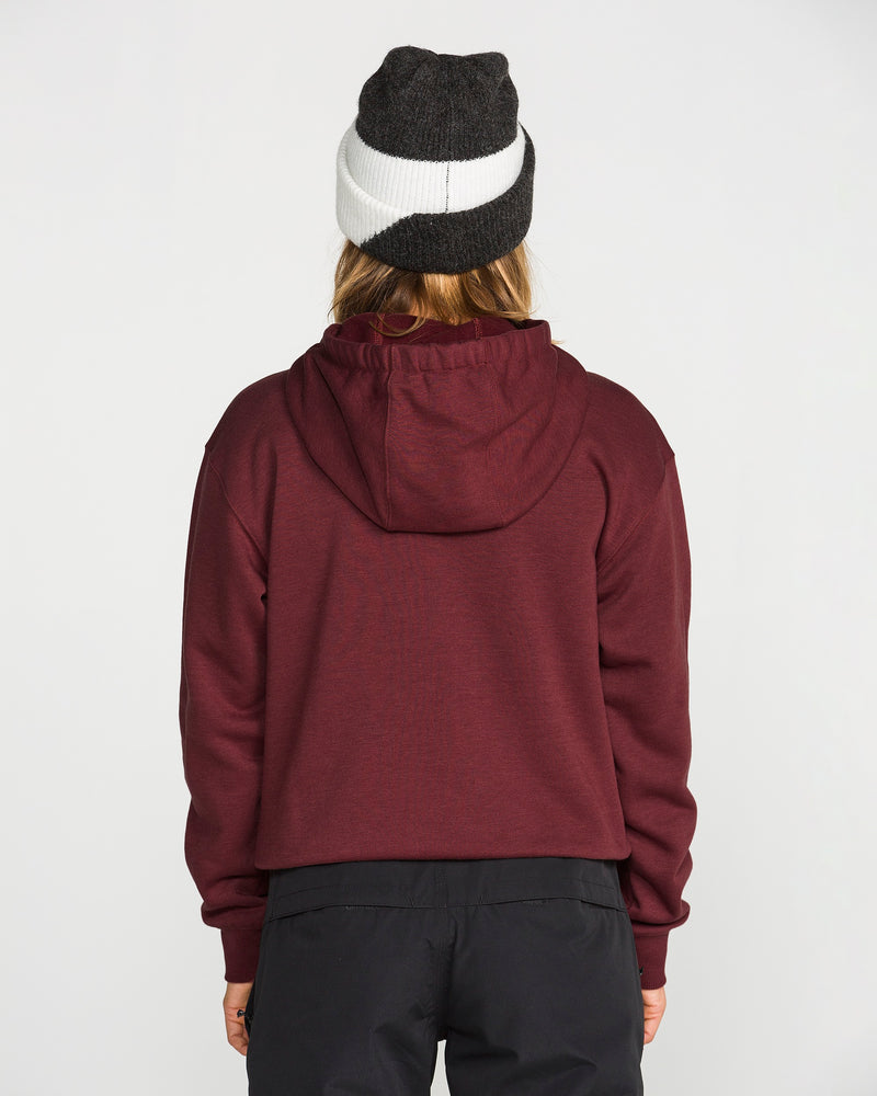 A person facing away, wearing the Volcom Women’s Alpine All Day Pullover in maroon with a funnel hood and handwarmer pocket, paired with black pants and a black and white beanie, stands against a plain white background.
