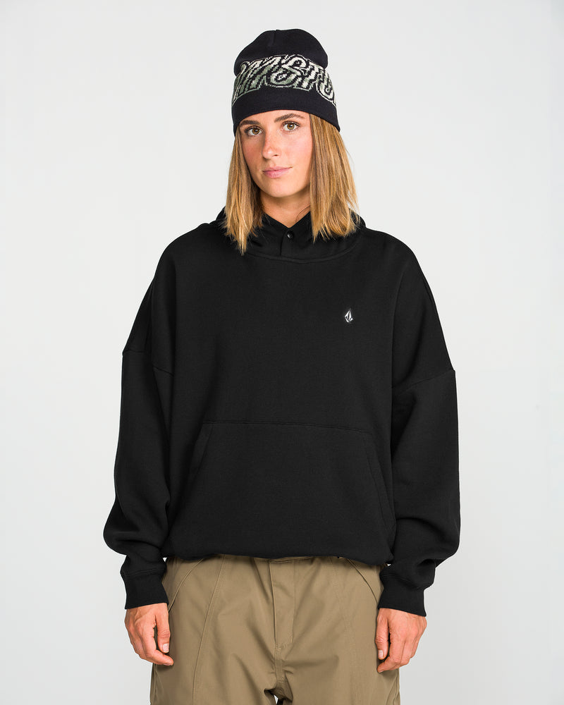 A person stands facing forward against a plain light background, wearing the Volcom Women’s Yourfav Baggy Pullover in black, paired with khaki pants and a black beanie with a white pattern.