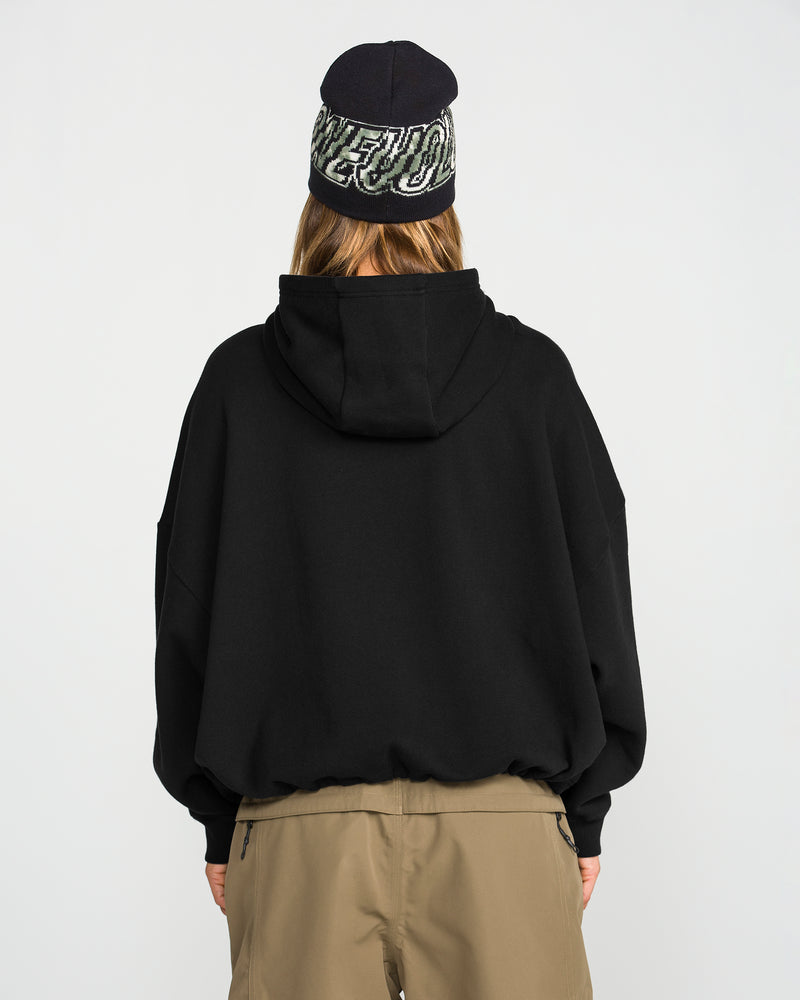 A person is shown from behind, wearing the Volcom Women’s Yourfav Baggy Pullover in black, paired with tan pants and a dark beanie featuring white lettering, standing against a plain white background.