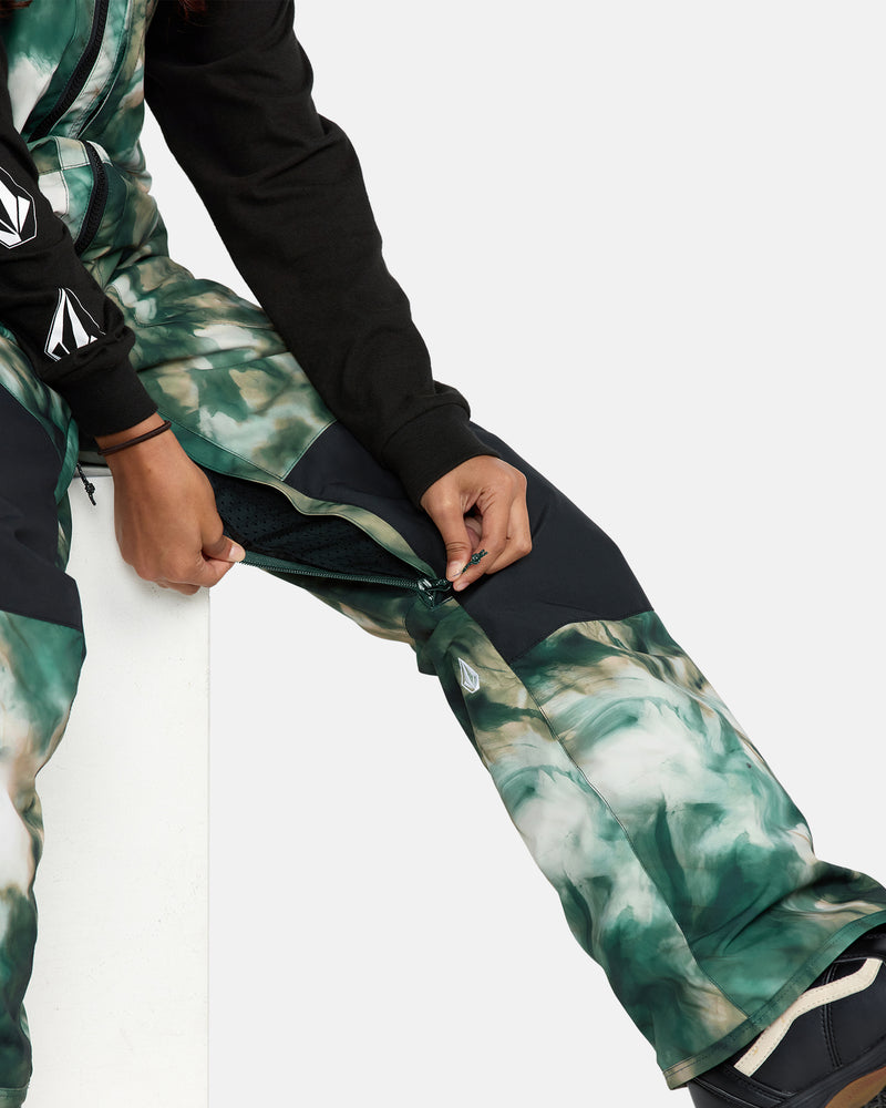 A person seated in a black long-sleeve shirt unzips a vent on their Kids' Barkley Insulated Bib Overall, revealing mesh fabric beneath the green and white patterned pants.