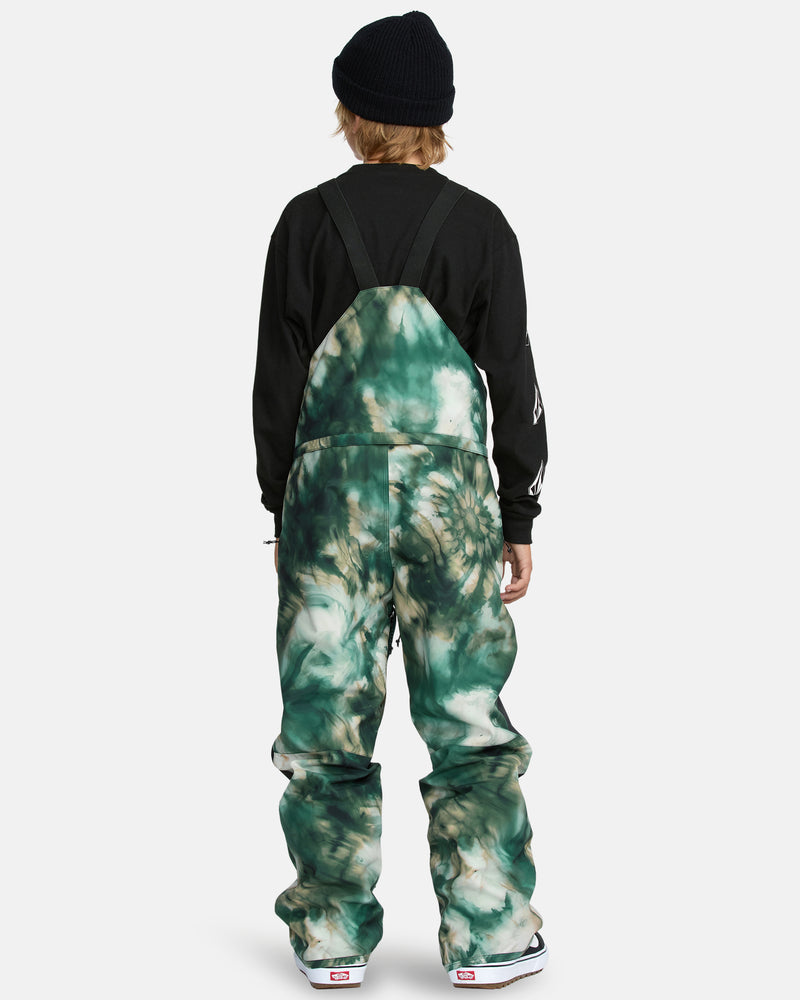 A person faces away from the camera wearing a black beanie, black long-sleeve shirt, and Kids' Barkley Insulated Bib Overall in green tie-dye with straps and loose fit, covering white shoes with red accents.