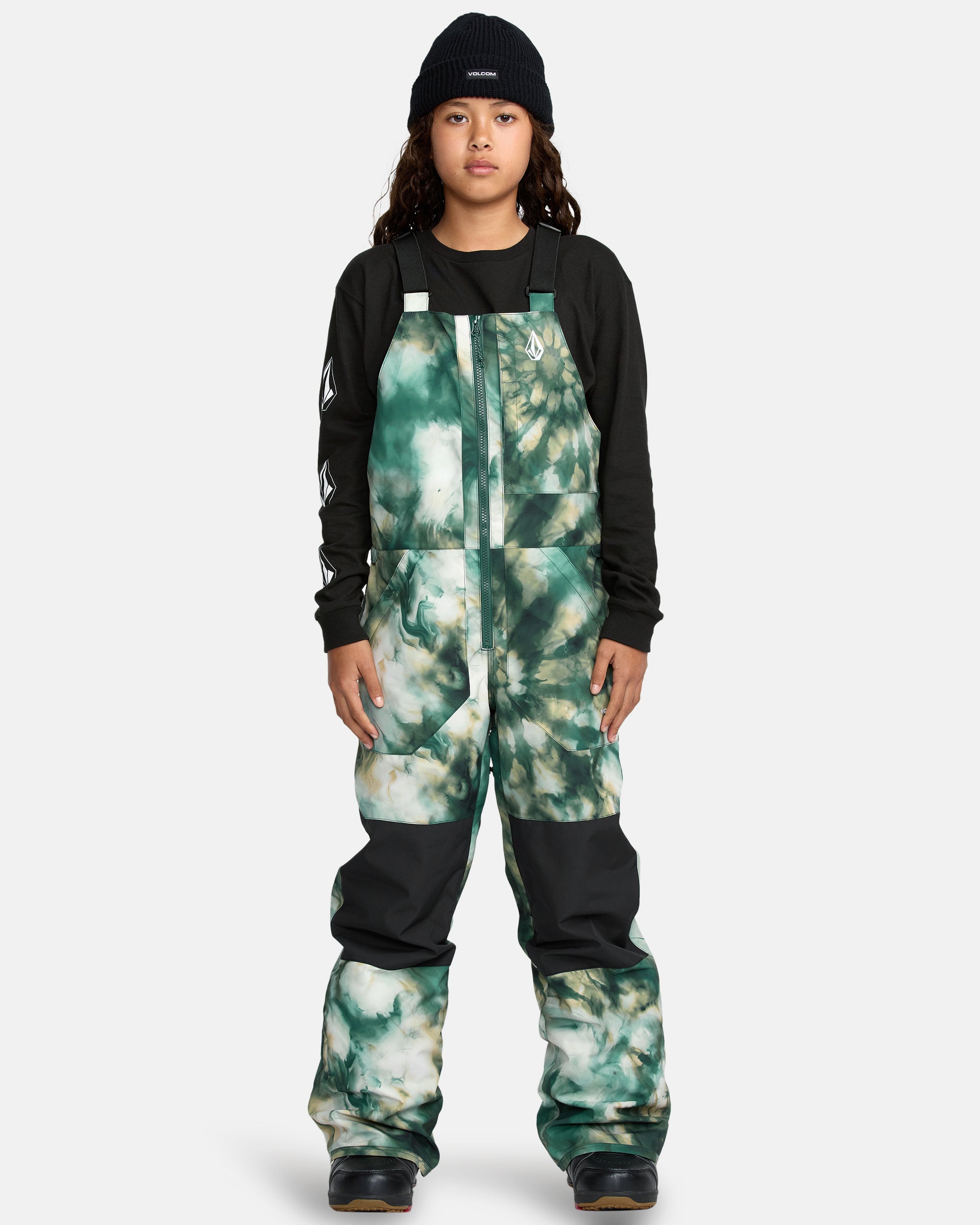A child models the Kids' Barkley Insulated Bib Overall in green-and-white tie-dye, paired with a black beanie and black long-sleeve shirt, standing against a plain light gray background.