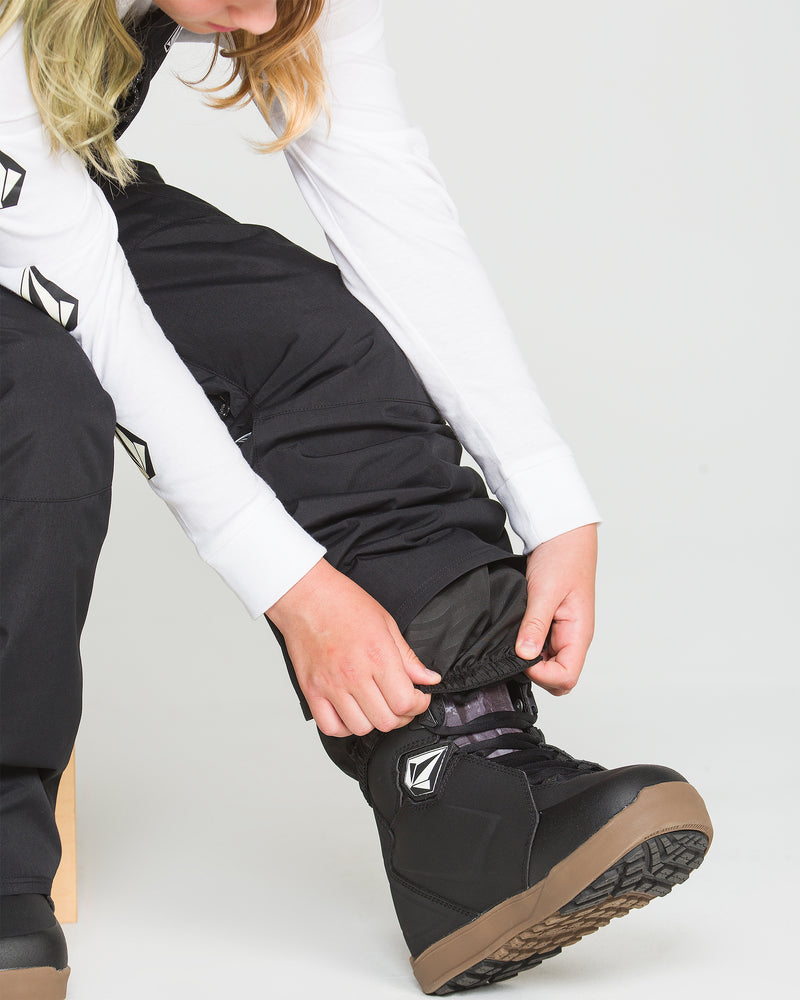 A person is wearing the Volcom Kids’ Youth Barkley Insulated Bib in black along with black snowboard boots, adjusting the pant leg over the boot. Only the lower half of their face and body is visible against a plain white background.
