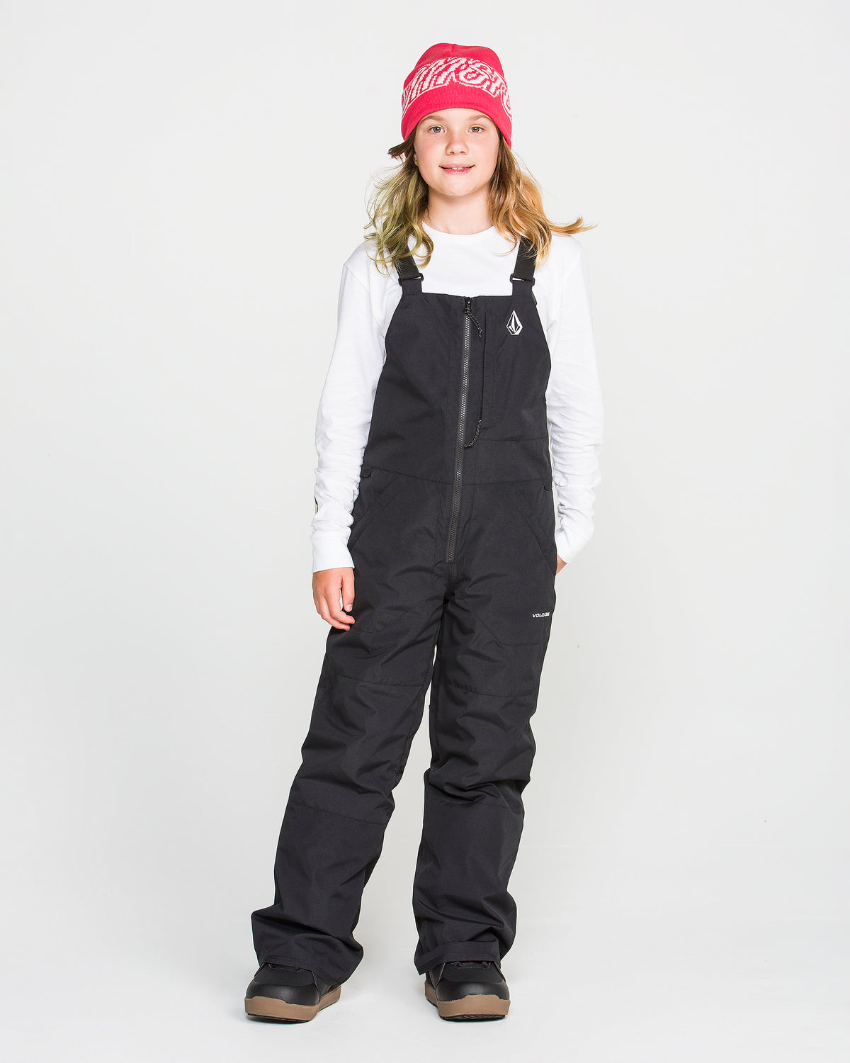 A young person wearing a pink beanie, white long-sleeve shirt, Volcom Kids’ Youth Barkley Insulated Bib in black with waterproof insulation, and brown winter boots stands facing forward against a plain white background.