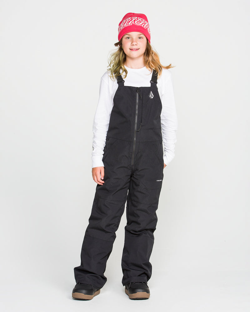 A young person wearing a pink beanie, white long-sleeve shirt, Volcom Kids’ Youth Barkley Insulated Bib in black with waterproof insulation, and brown winter boots stands facing forward against a plain white background.