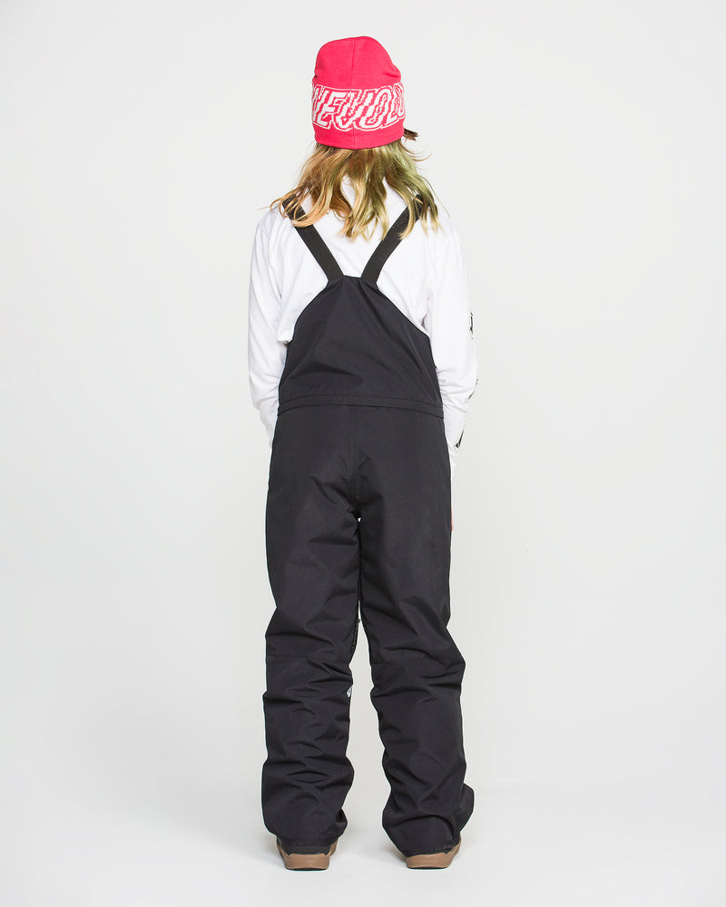 A person stands facing away, wearing a pink beanie, white long-sleeve shirt, and the Volcom Kids’ Youth Barkley Insulated Bib—black snow overalls with straps featuring waterproof insulation—against a plain white background.