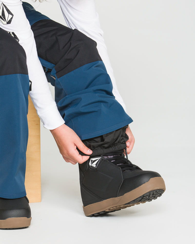 A person wearing the Volcom Kids’ Youth Barkley Insulated Bib snow pants in blue and black with recycled insulation and a white long-sleeve shirt fastens a black snowboard boot while sitting on a light wooden bench.