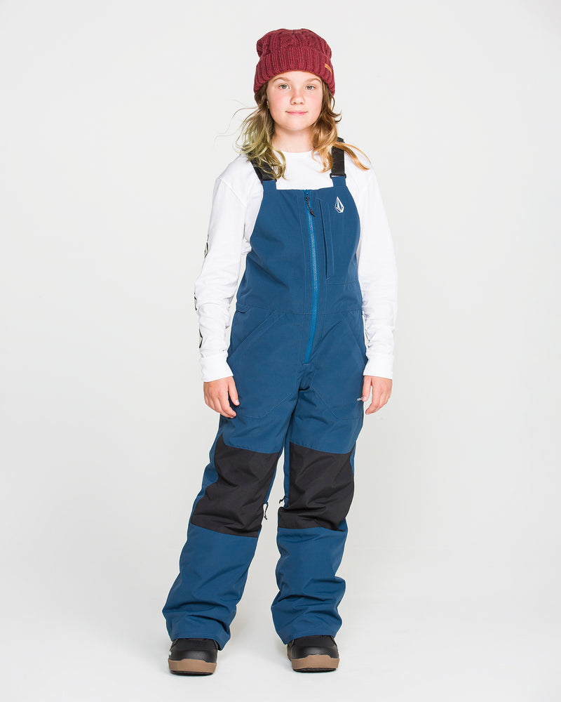 A child stands against a white background, wearing a red knit beanie, a white long-sleeve shirt, and Volcom Kids’ Youth Barkley Insulated Bib snow overalls in blue with black knee patches and brown boots. The overalls offer recycled insulation and waterproof breathability for all-day comfort and warmth.