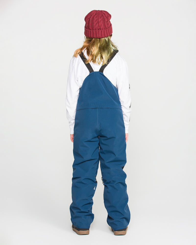 A person is shown from behind, wearing a red knit hat, a white long-sleeve shirt, and blue Volcom Kids’ Youth Barkley Insulated Bib snow overalls featuring waterproof breathability with recycled insulation, paired with brown boots against a plain white background.