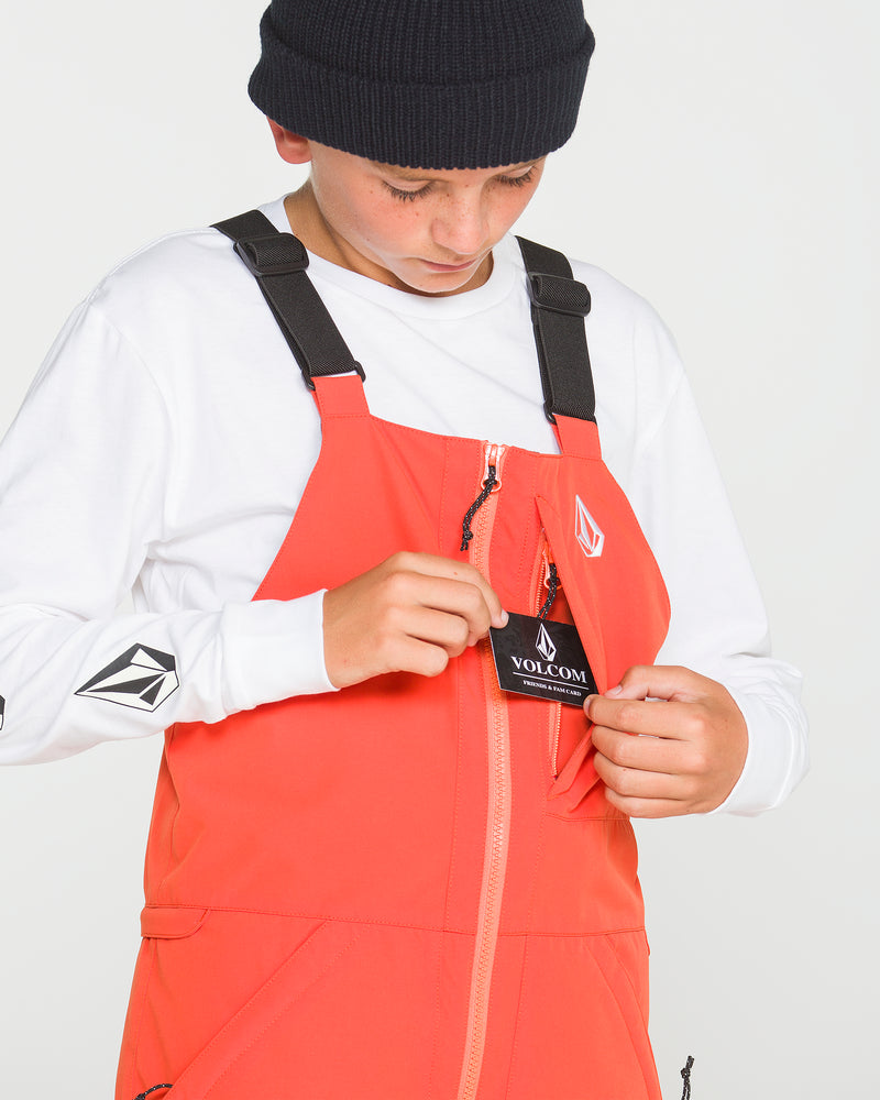 A person in a black beanie and white long-sleeve shirt opens the chest pocket of bright orange Volcom Kids’ Youth Barkley Insulated Bib overalls to reveal the Volcom tag, looking down at it and highlighting the style’s waterproof breathability.
