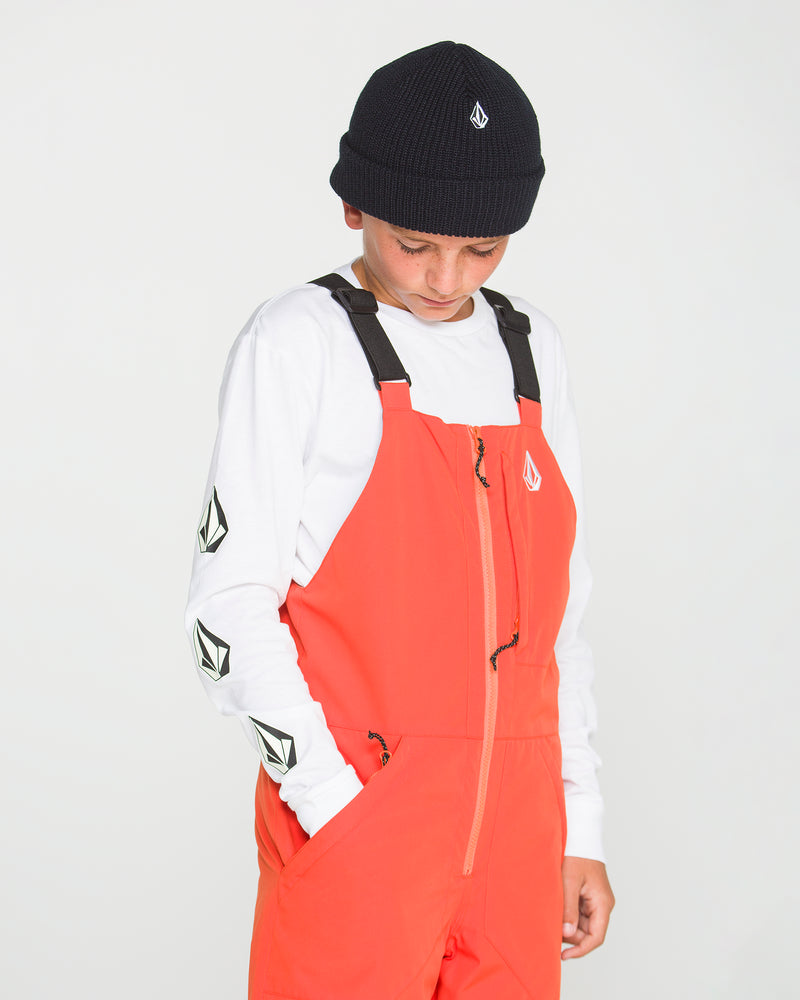 A person wearing a black beanie, a white long sleeve shirt with black logos on the sleeve, and Volcom’s Kids’ Youth Barkley Insulated Bib in bright orange—with recycled insulation—looks down with hands in pockets against a plain white background.