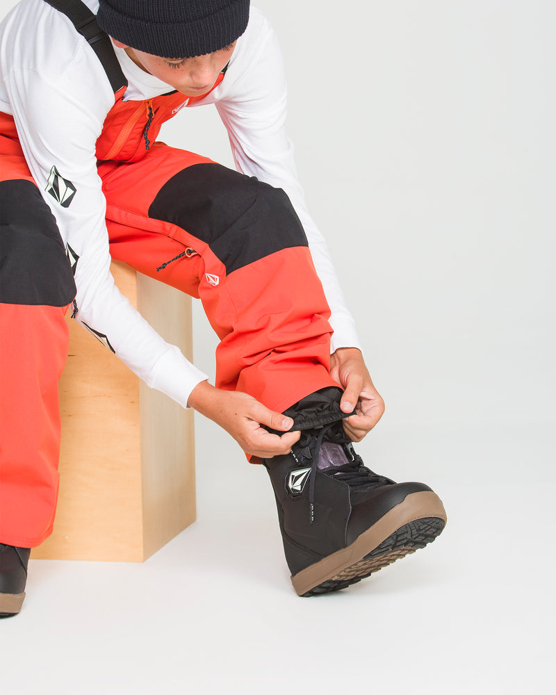 A person wearing the Volcom Kids’ Youth Barkley Insulated Bib in bright orange with recycled insulation and a black beanie sits on a wooden box, adjusting black snow boots over their pants against a plain white background.