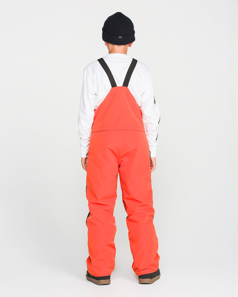 A person stands with their back to the camera, wearing a black beanie, a white long-sleeve shirt, and bright orange Volcom Kids’ Youth Barkley Insulated Bib overalls with black straps—featuring waterproof breathability and recycled insulation—set against a plain white background.