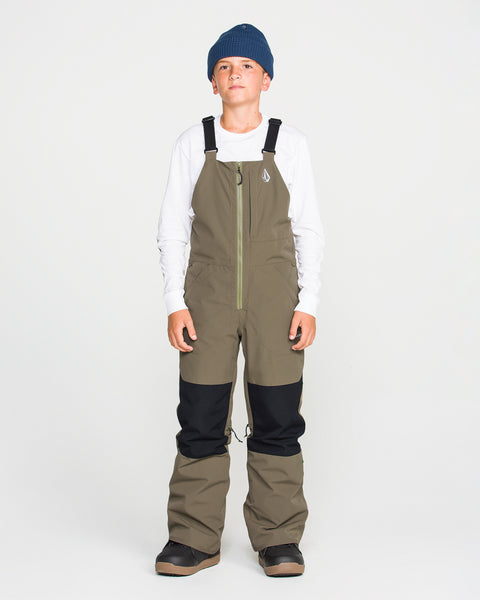 A boy wearing a blue beanie, white long-sleeve shirt, and Volcom Kids’ Youth Barkley Insulated Bib snow pants in olive green with black knee patches stands facing forward against a plain white background. The bib features waterproof insulation and Grow-Tech for lasting comfort and warmth.
