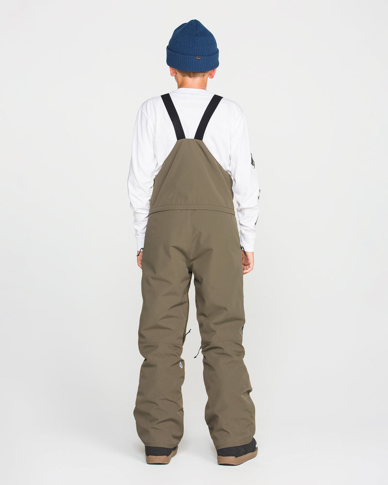 Person standing with back to camera, wearing a blue beanie, white long-sleeve shirt, and Volcom Kids’ Youth Barkley Insulated Bib snow overalls in olive green with black straps, featuring waterproof insulation and Grow-Tech technology, against a plain white background.