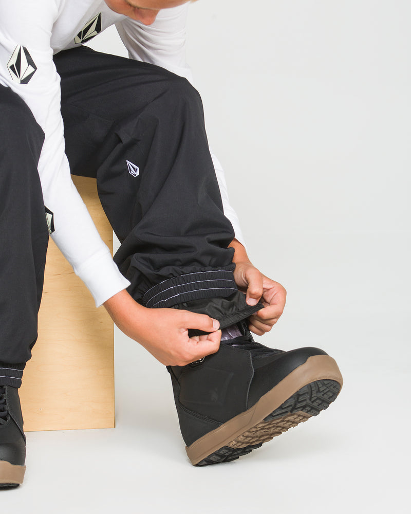 A person sits on a wooden box, wearing the Volcom Kids’ Youth Chaos Insulated Pants with 10K/10K protection and boots, adjusting the pant cuff over one boot. These pants feature recycled insulation for warmth. The background is plain white.