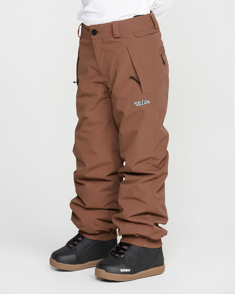 A child wearing Volcom Kids’ Youth Chaos Insulated Pants in brown, featuring recycled insulation and V-Science 2-Layer technology with the VOLCOM logo, paired with a white shirt and black winter boots, stands against a plain white background.