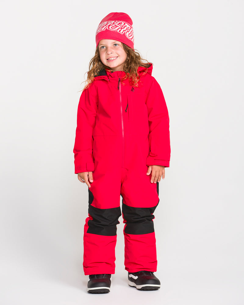 A young child stands smiling against a plain white background, wearing the Volcom Kids’ Kid’s Little Flurry One Piece snowsuit, a red hat with white writing, black gloves, and winter boots.