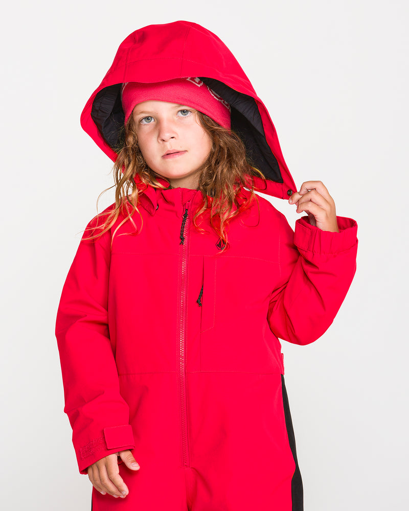 A young child with long hair wears the Volcom Kids’ Little Flurry One Piece in bright red, along with a matching hat, holding the hood with one hand while standing against a plain white background. This insulated snowsuit boasts 10K/10K protection and is crafted from recycled V-Science fabric for excellent warmth and sustainability.