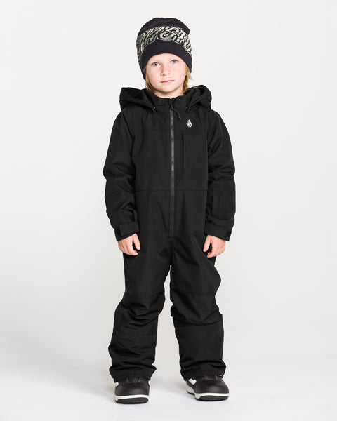 A child models the Volcom Kids’ Little Little Flurry One Piece Snow Suit in black, paired with a black and white knit hat and dark shoes, standing facing forward against a plain white background.