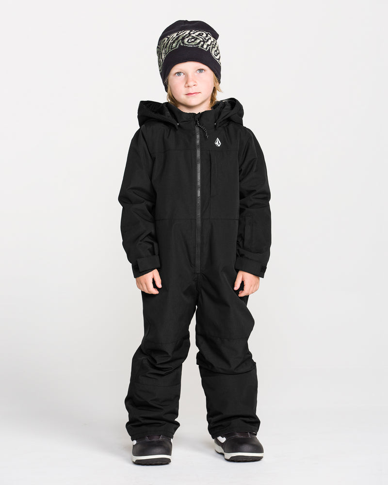 A child models the Volcom Kids’ Little Little Flurry One Piece Snow Suit in black, paired with a black and white knit hat and dark shoes, standing facing forward against a plain white background.