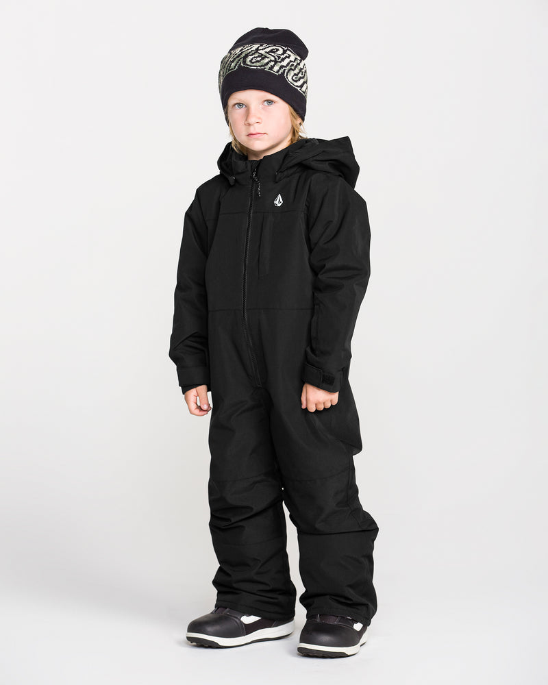 A young child stands against a plain white background wearing the Volcom Kids’ Little Little Flurry One Piece Snow Suit, paired with black winter boots and a black beanie with white lettering.