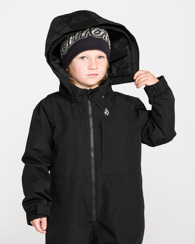A young child wearing the Volcom Kids’ Little Little Flurry One Piece Snow Suit and a patterned beanie looks at the camera, holding the hood with one hand against a plain light background.