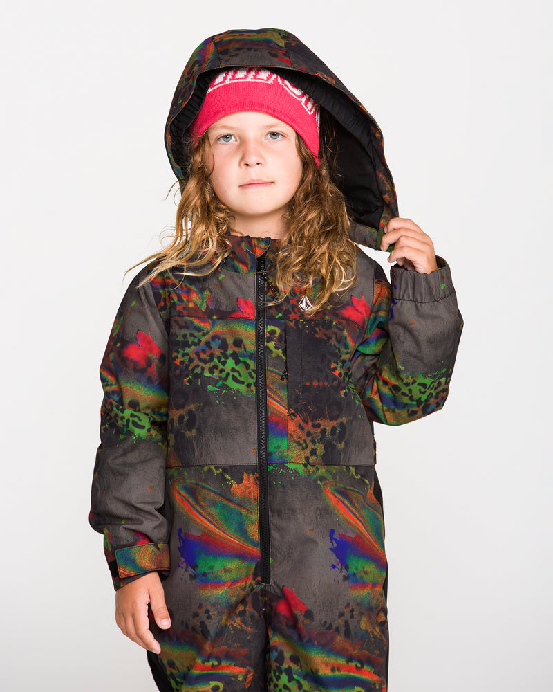 A young child with long hair wears the Volcom Kids’ Little Flurry One Piece—a colorful, patterned insulated suit featuring a helmet-compatible removable hood—and a red beanie, standing against a plain white background while holding up the hood with one hand.