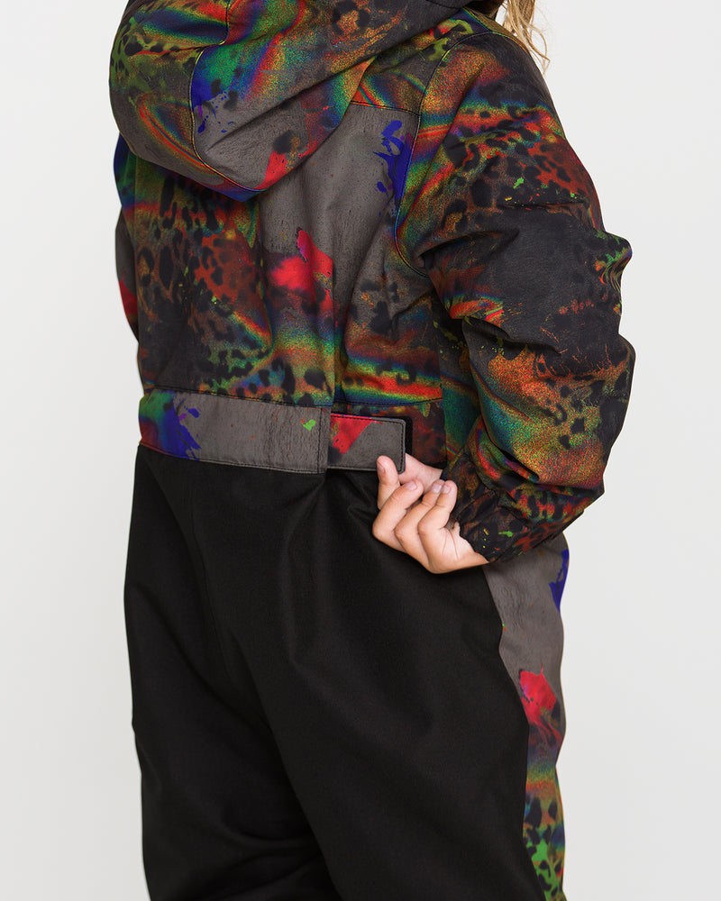 A child wearing the Volcom Kids’ Little Flurry One Piece, featuring a colorful abstract pattern, warm insulation, and a helmet-compatible removable hood, adjusts a side strap. The photo is taken from behind, showing the back and part of the side.