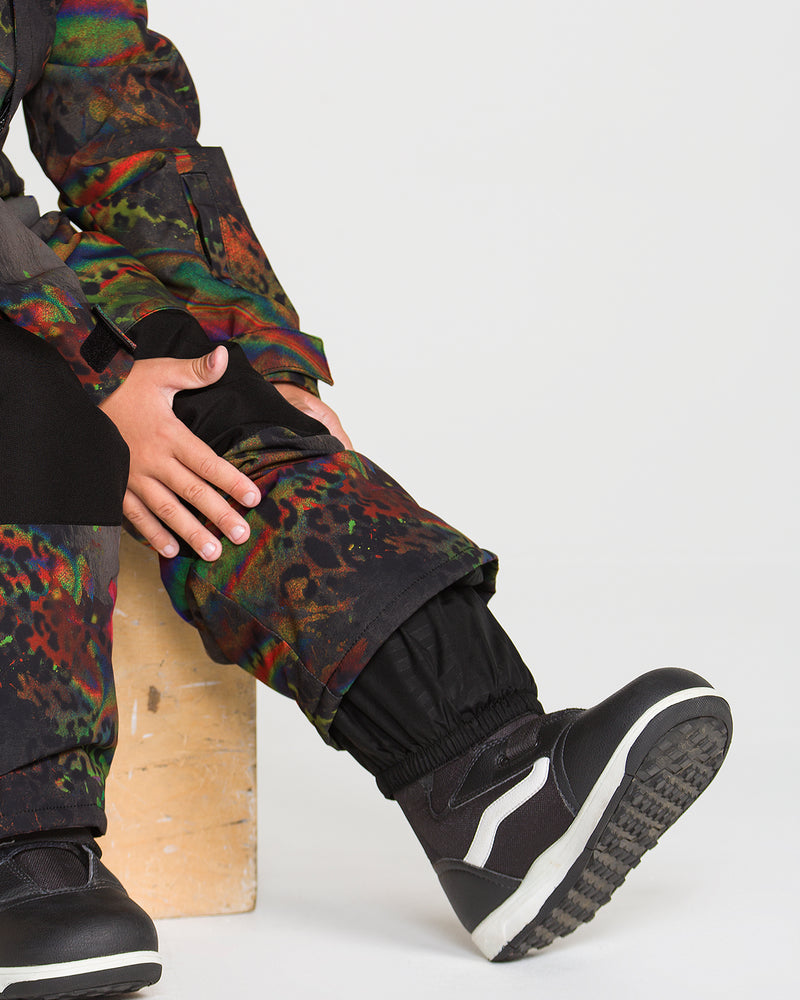 A child wearing the Volcom Kids’ Little Flurry One Piece with colorful patterns sits on a wooden block, black sneakers on their feet and hands resting on their leg, against a plain white background.