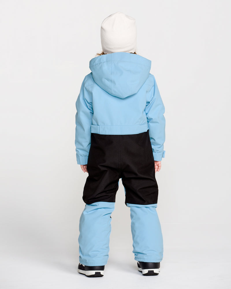 A child is seen from behind against a plain white background, wearing the Volcom Kids’ Little Flurry One Piece snowsuit in light blue and black, which features recycled V-Science fabric for enhanced sustainability, paired with a hood and a white beanie.