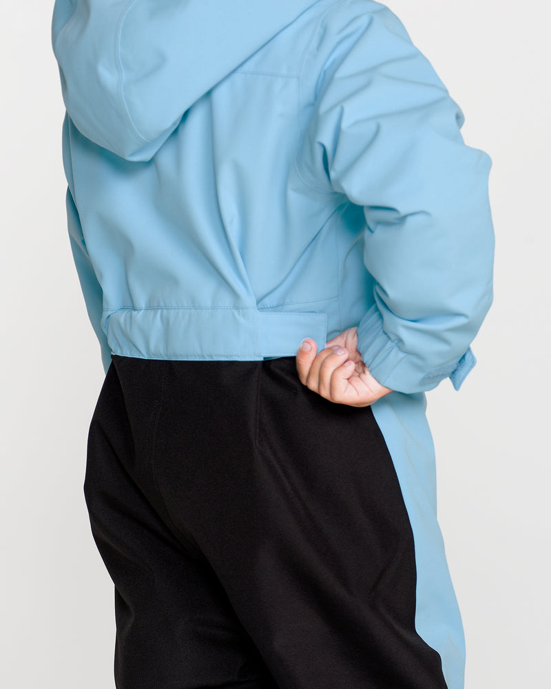 A child wearing the Volcom Kids’ Kid’s Little Flurry One Piece in light blue—crafted from recycled V-Science fabric—is pictured from behind, standing with one hand on their hip.