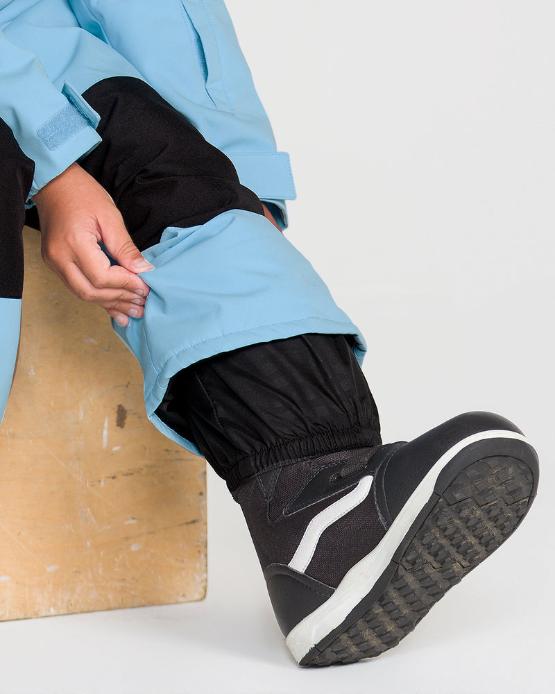 A person sits on a wooden box against a plain white background, wearing the Volcom Kids’ Little Flurry One Piece in light blue—crafted with recycled V-Science fabric—and black-and-white winter boots with 10K/10K protection. They pull up the leg of their one piece to reveal the boot.