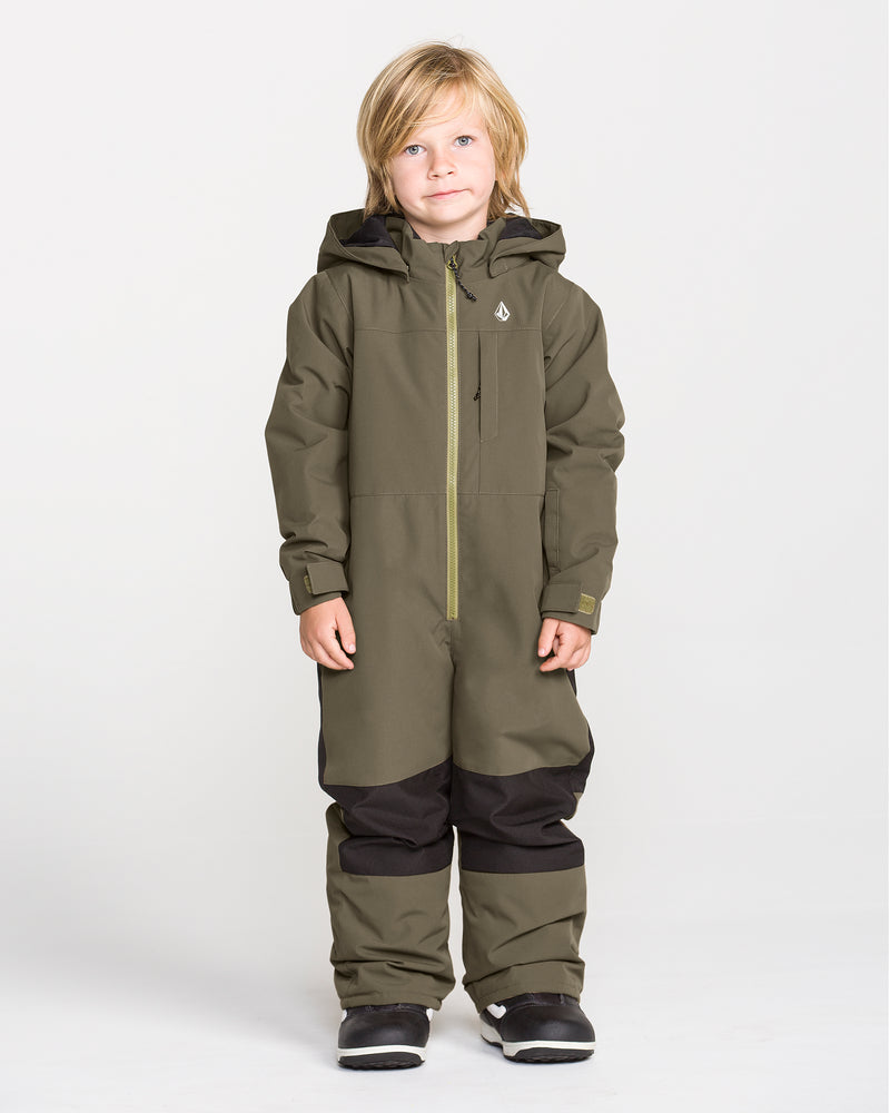 A young child with blonde hair stands against a plain white background, dressed in the Volcom Kids’ Little Flurry One Piece snowboard suit in olive green, which features black knee patches and black boots. This insulated snowsuit is crafted from recycled V-Science materials for warmth and sustainability.