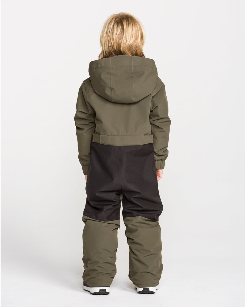 A child with long blonde hair stands facing away, wearing the Volcom Kids’ Kid’s Little Flurry One Piece snowboard suit in olive green and black, crafted from recycled V-Science materials, paired with white shoes against a plain white background.