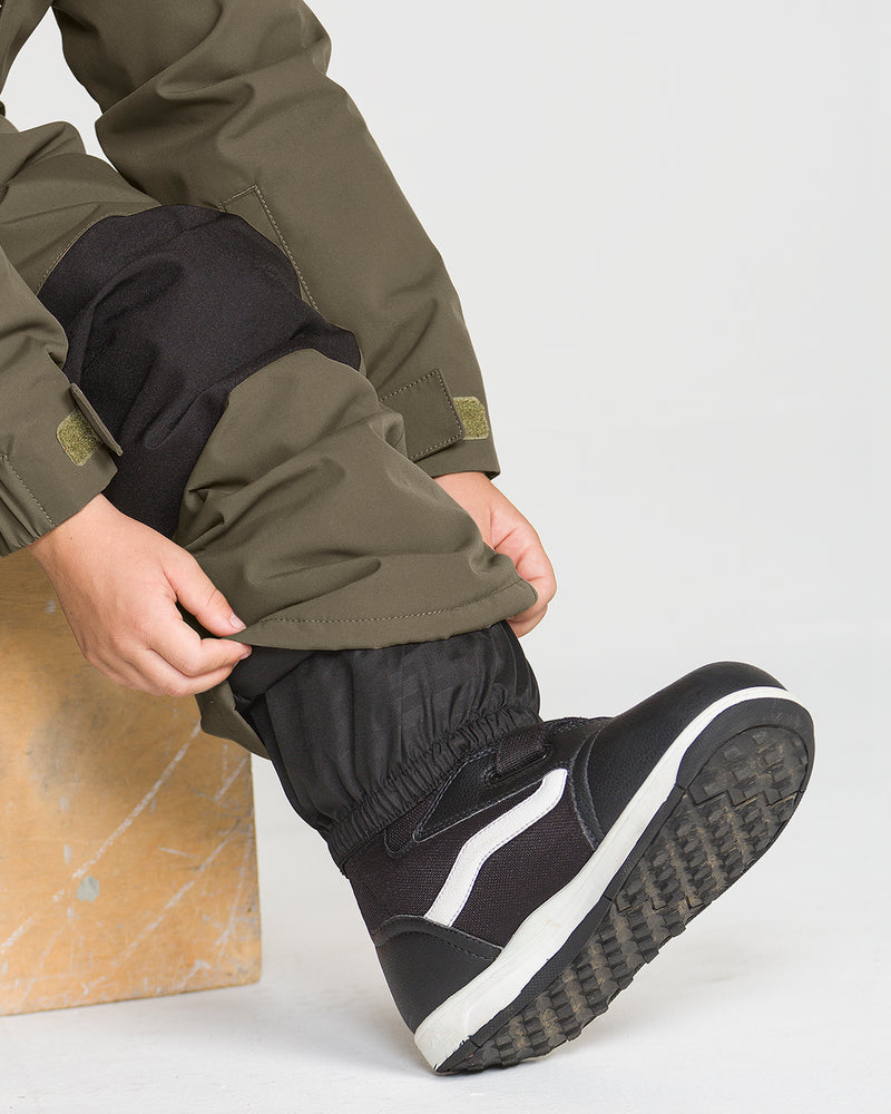 A child wearing olive green Volcom Kids’ Little Flurry One Piece snow suit and black boots adjusts the cuff of their insulated outfit while sitting on a wooden block.