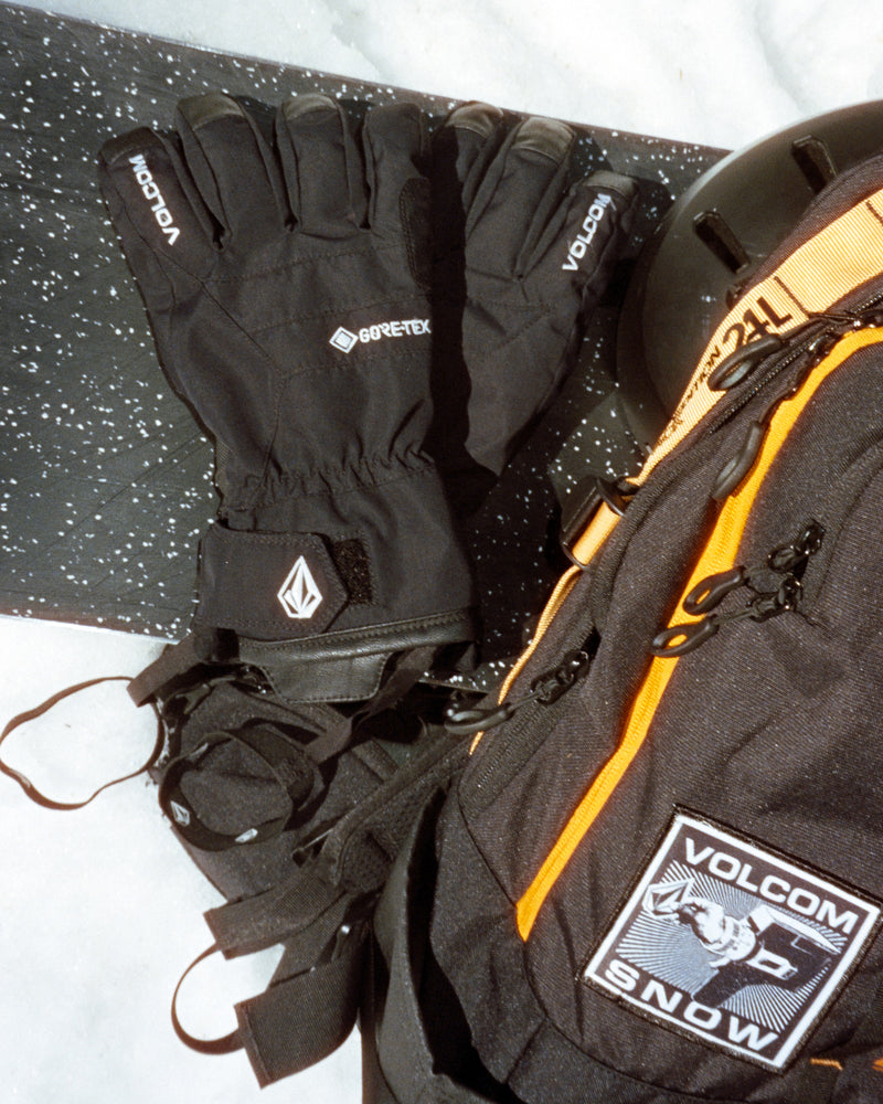 Black Men’s Cp2 Gore-Tex Gloves with wrist straps resting on a snowboard next to a black and orange backpack.