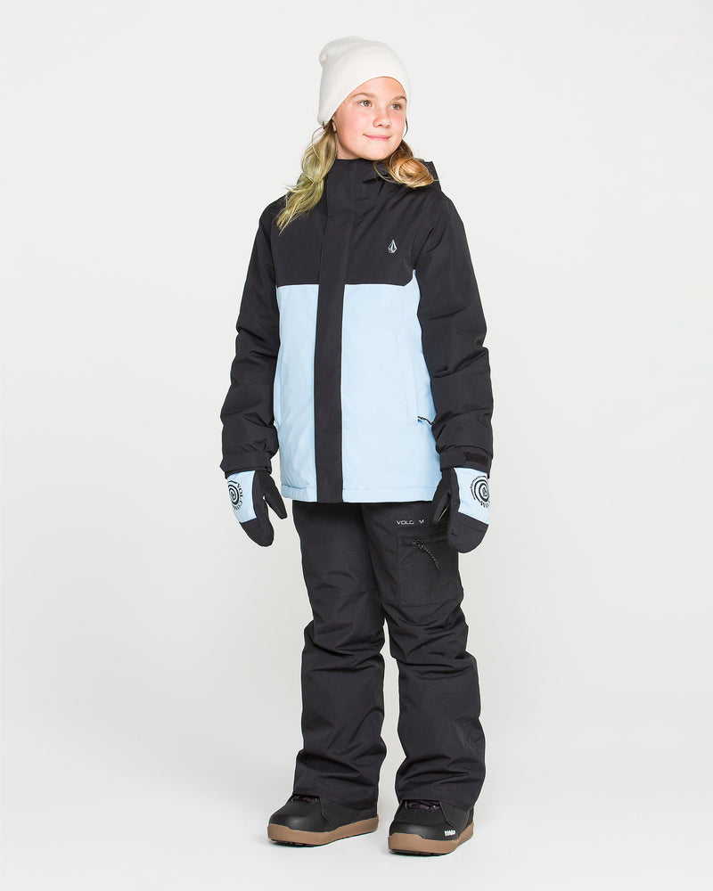 A child wearing the Volcom Kids’ Sass’N’Frass Insulated Jacket in black and light blue, paired with black snow pants, gloves, brown boots, and a white beanie stands against a plain white background, looking slightly to the side and smiling.