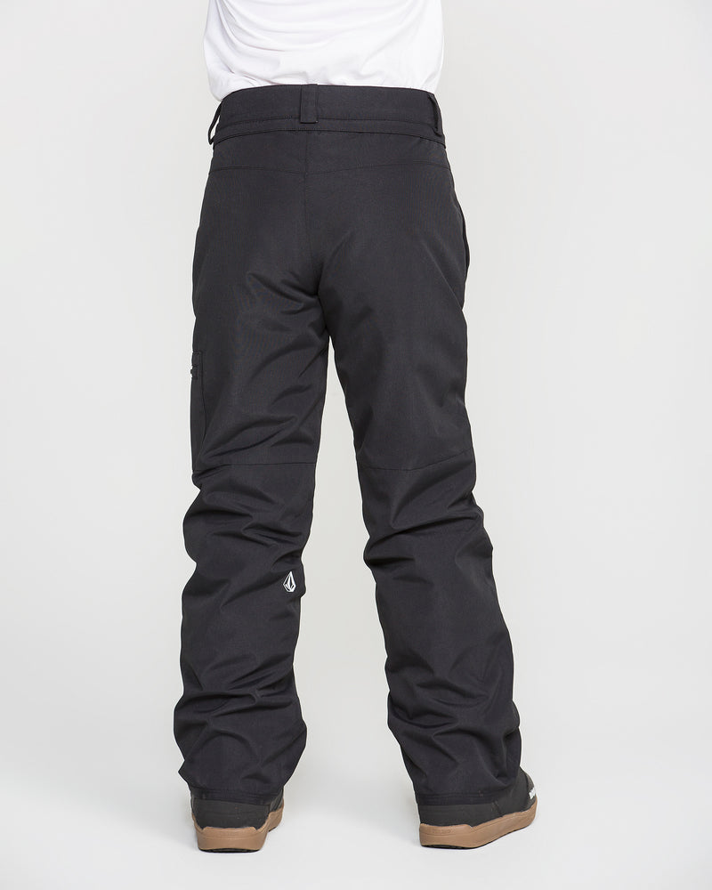 A child is seen from behind, standing against a plain white background and wearing Volcom Kids’ Frostique Insulated Pants in black. These pants feature an adjustable waist, relaxed fit, visible seams, and are designed by Volcom for comfort and protection. The child pairs the snow pants with brown boots.