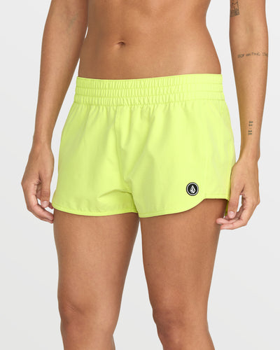 A person wearing Women's Simply Solid Boardshorts in neon yellow, made from recycled polyester with an elastic waistband and a small black and white logo on the left leg, stands against a plain light background.