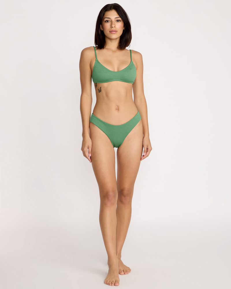 A woman stands barefoot against a light background, wearing the Volcom Women's Pucker Up Scoop Crop Bikini Top in green with thin straps and textured recycled nylon fabric. She has straight dark hair, a neutral expression, and a left ribcage tattoo.