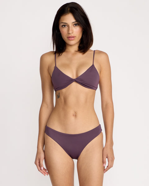 A woman with straight, dark hair faces forward in a mauve Volcom Women's Simply Seamless Vneck bikini with thin straps. She has a small tattoo on her right ribcage and a neutral expression. The background is plain white.