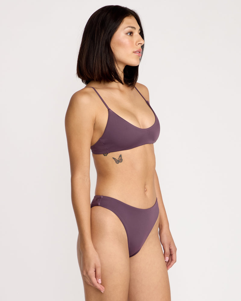A woman with straight dark hair, wearing the Volcom Women's Simply Seamless Skimpy mauve bikini, stands in profile against a plain white background. A small butterfly tattoo is visible on her left side below her ribs.