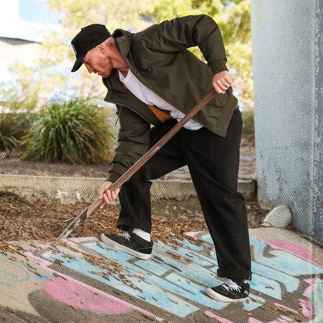 A man wearing an olive green utility jacket, black pants, and a black cap is bent over using a long-handled brush to clean graffiti off of a concrete surface outdoors.