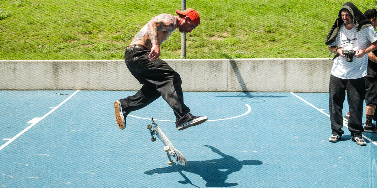 A skateboarder in a red cap, sleeveless shirt, and black pants jumps mid-air on a blue outdoor court, with a skateboard beneath the board and two onlookers standing nearby against a grassy background.