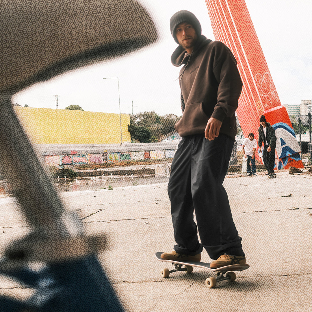 A person wearing a brown hoodie, dark pants, and a black beanie stands on a skateboard in an outdoor urban setting with graffiti walls and a large red structure in the background.