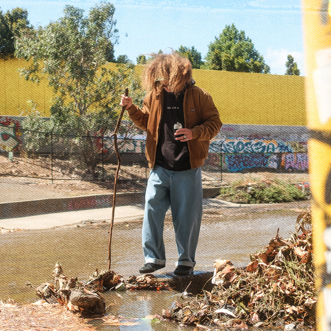 A person standing outdoors in a muddy area near a fence, wearing a brown jacket, black shirt, and light blue jeans, holding a long stick and a small bottle. Trees, a yellow wall, and graffiti are visible in the background.