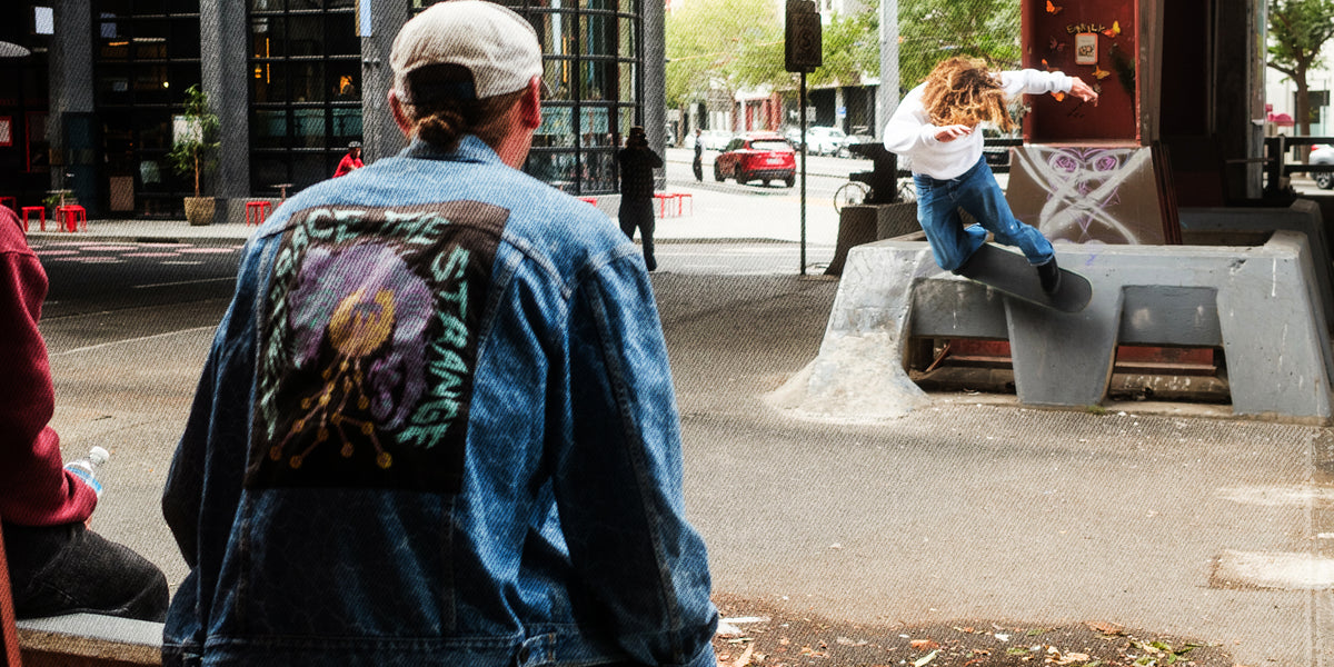 A person wearing a blue denim jacket with a large colorful graphic print on the back stands in an urban street setting, facing a skateboarder performing a trick on a concrete ramp.