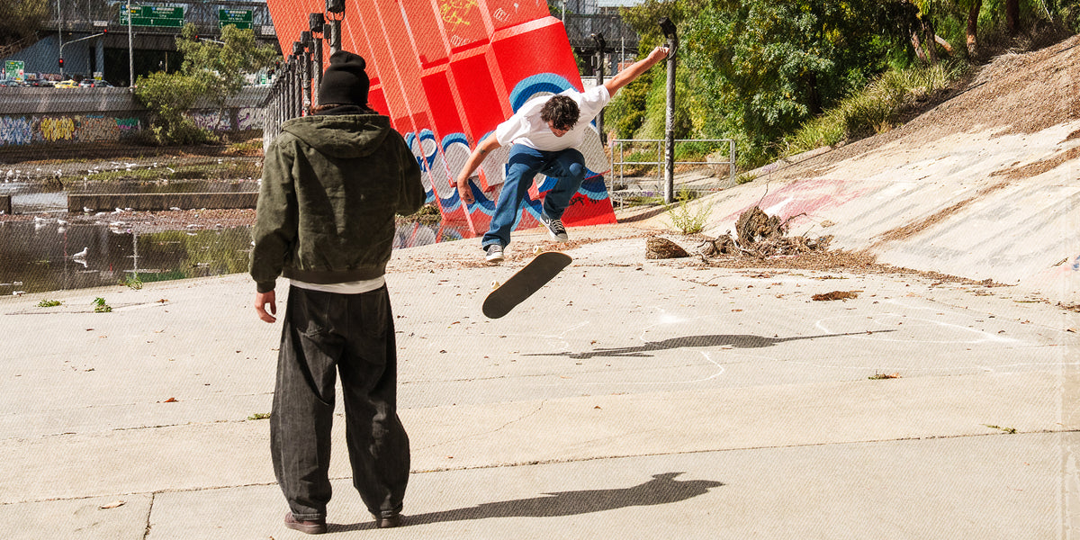 A skateboarder in a white t-shirt and blue jeans performs a trick mid-air in an outdoor concrete skatepark, with another person standing in the foreground and a large red graffiti-covered structure in the background.